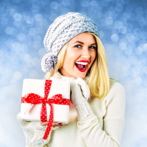 Smiling female wearing sweater, mittens, and winter hat holding wrapped present with joy