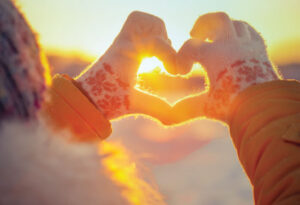Two hands in winter gloves make a heart shape with sun shining through
