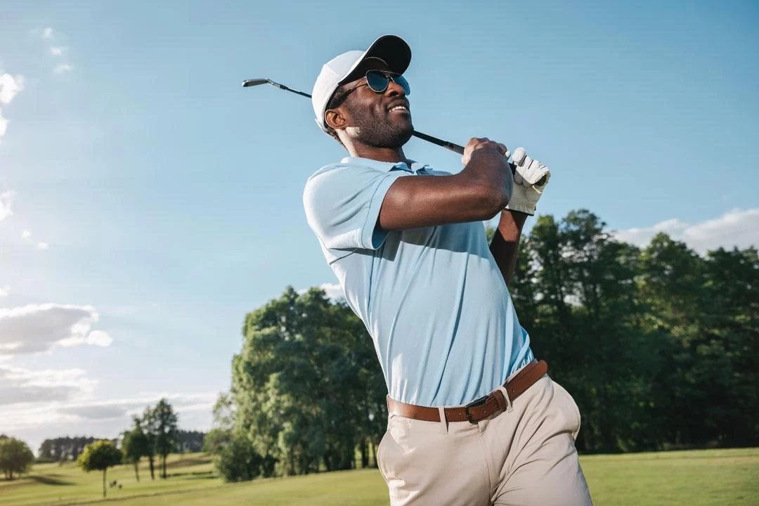 A black golfer swings at the ball on a golf course during a sunny day.
