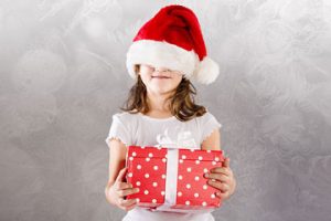 Girl holding a gift with a Santa hat covering her eyes.