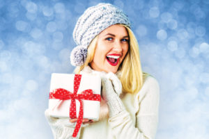 Smiling female wearing sweater, mittens, and winter hat holding wrapped present with joy