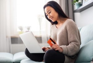 Young asian woman sitting on couch shopping online with laptop; debit card in hand