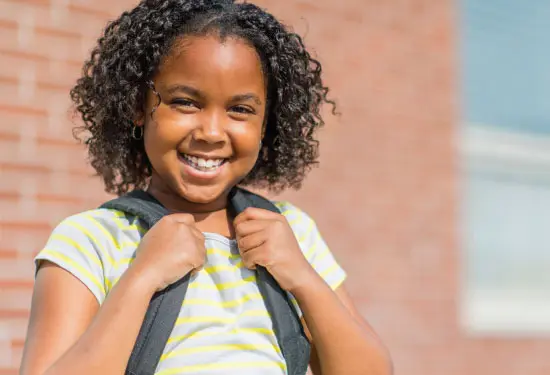A young girl smiles holding her backpack straps.